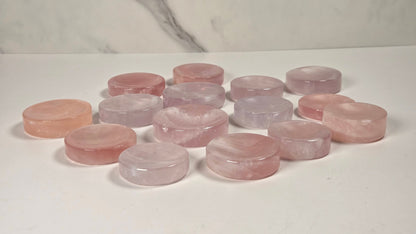 A group of round, polished pink and pale lavender stone discs, likely rose quartz or similar gemstone slices, arranged on a white surface with a light gray marble-patterned background.