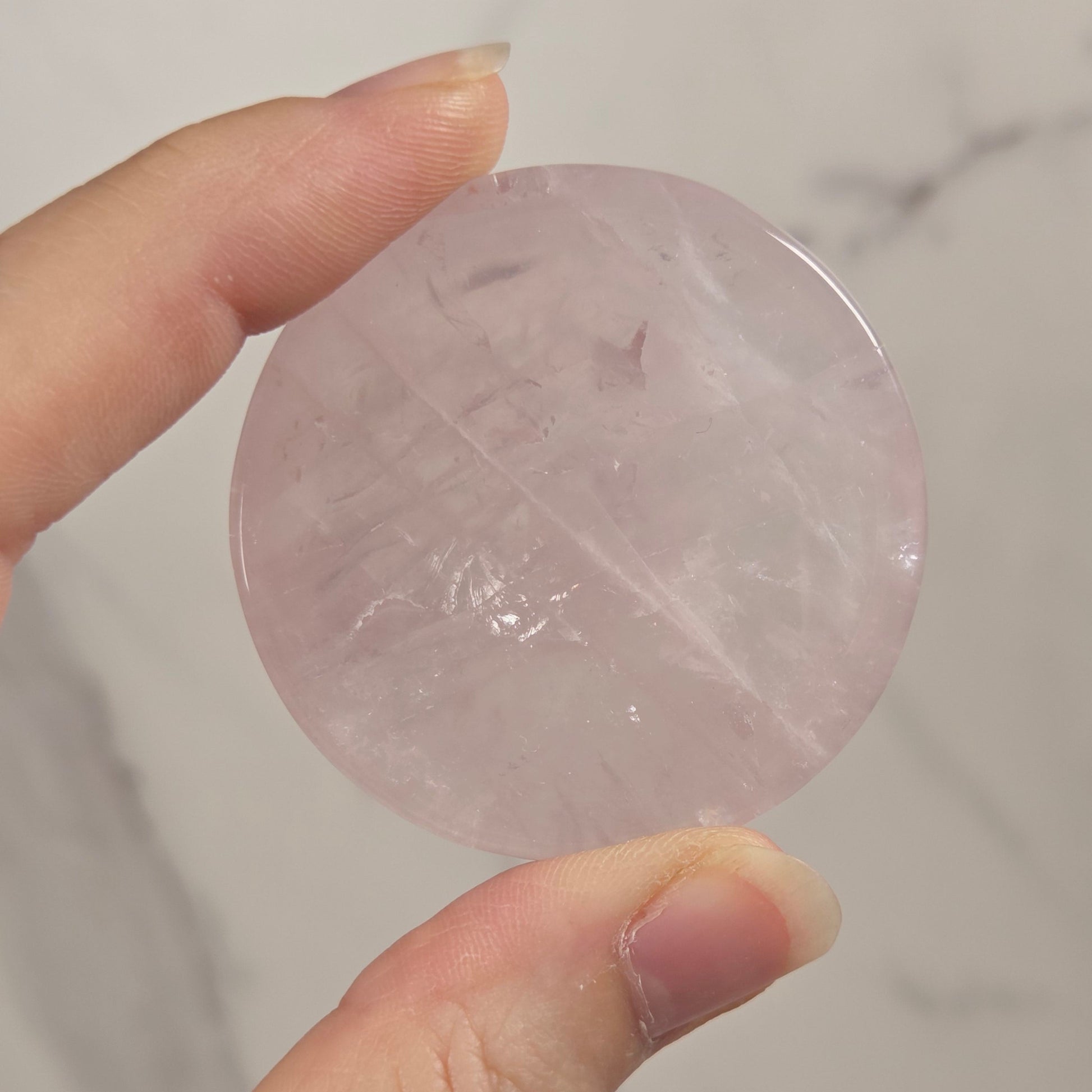 A hand holding a round, polished pink stone or crystal pendant against a light gray marble-patterned background.