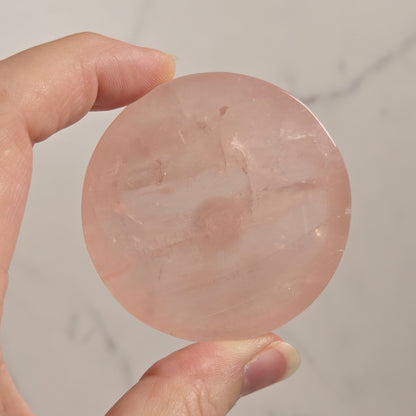 A hand holding a round, polished pink stone or crystal against a light marble-like background.