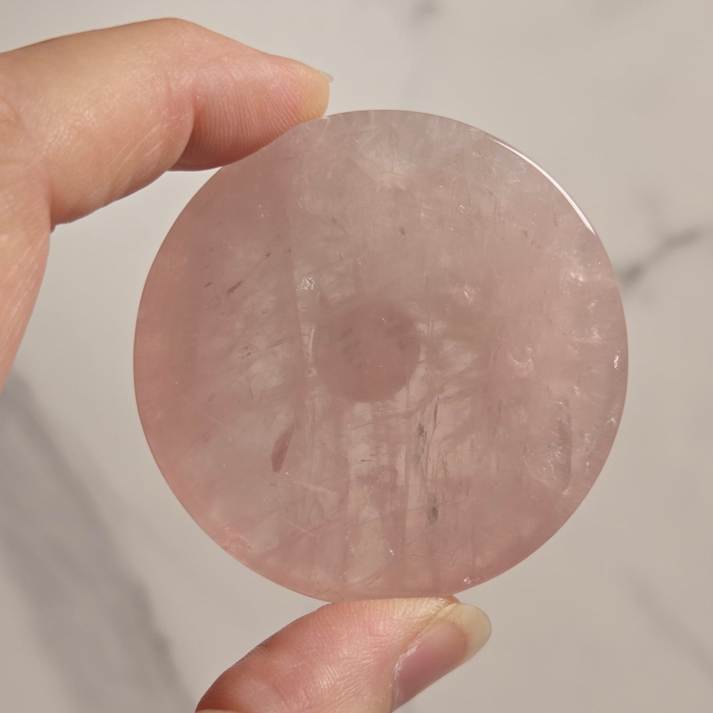 A hand holding a round, flat pink stone disc against a light marble-patterned background.