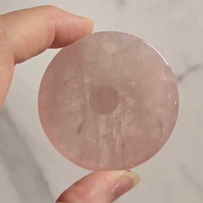 A hand holding a round, flat pink stone disc against a light marble-patterned background.