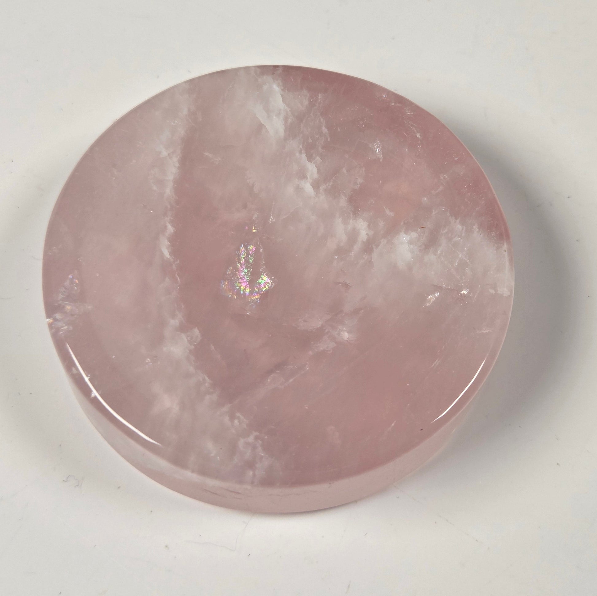A round, polished pink stone slab, likely rose quartz, with natural cloudy veining and a small iridescent inclusions spot near the center, resting on a plain off-white surface.