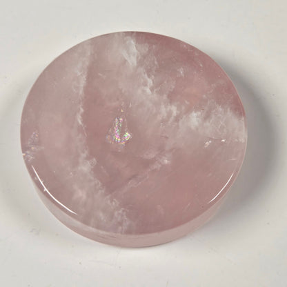 A round, polished pink stone slab, likely rose quartz, with natural cloudy veining and a small iridescent inclusions spot near the center, resting on a plain off-white surface.