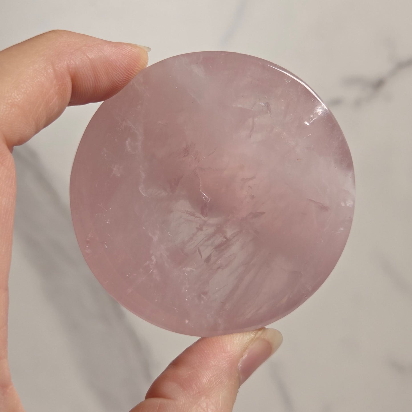 A hand holding a round, polished pink stone or crystal against a light marble-like background.