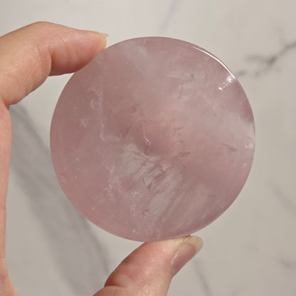 A hand holding a round, polished pink stone or crystal against a light marble-like background.