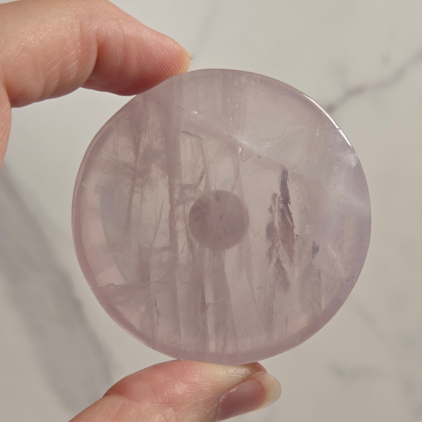 A hand holding a round, polished pink stone or crystal with natural inclusions, set against a light marble background.