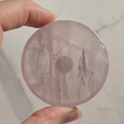 A hand holding a round, polished pink stone or crystal with natural inclusions, set against a light marble background.