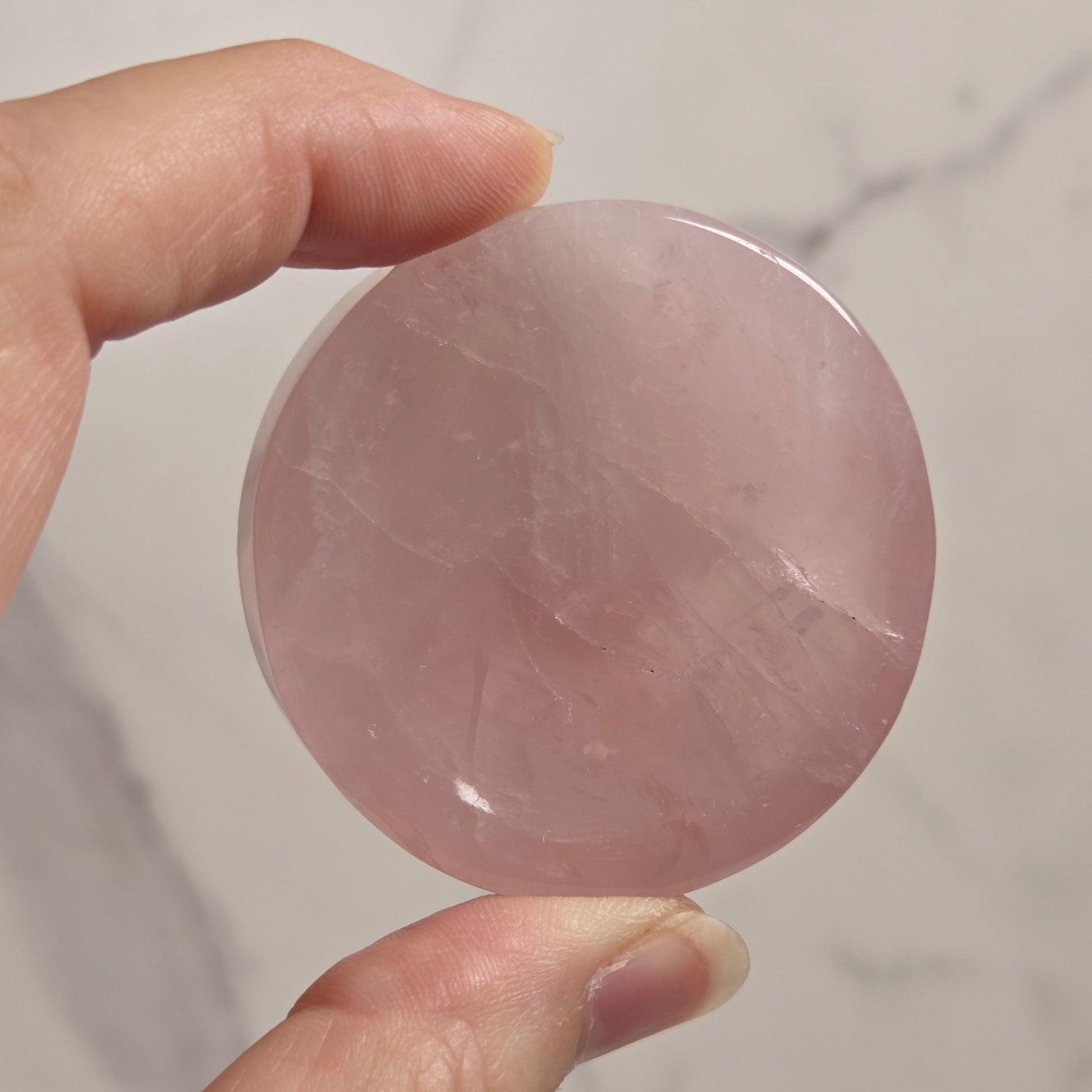 A hand holding a smooth, round pink stone disc against a light marble-patterned background.