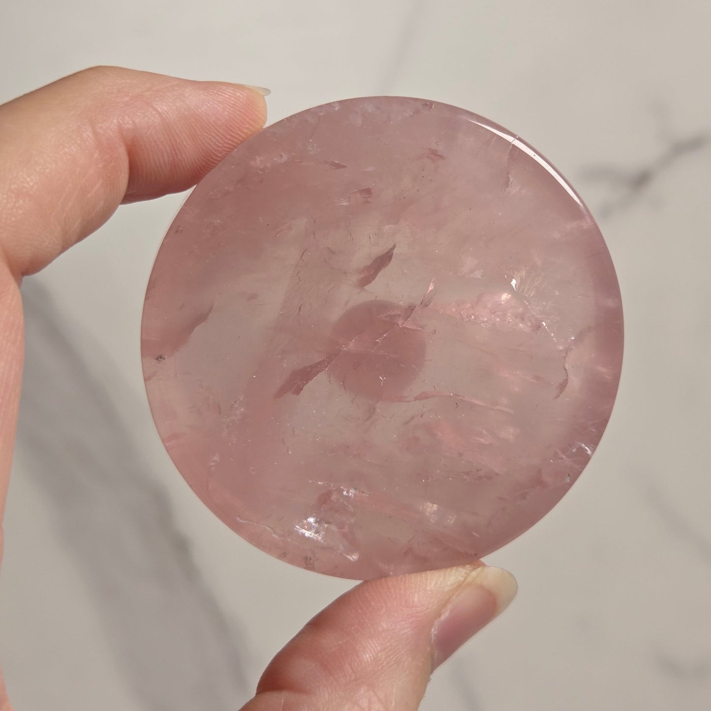 A hand holding a round, polished pink stone or crystal against a light marble-patterned background.
