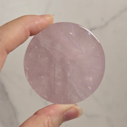 A hand holding a round, polished pink stone or crystal against a light marble-like background.