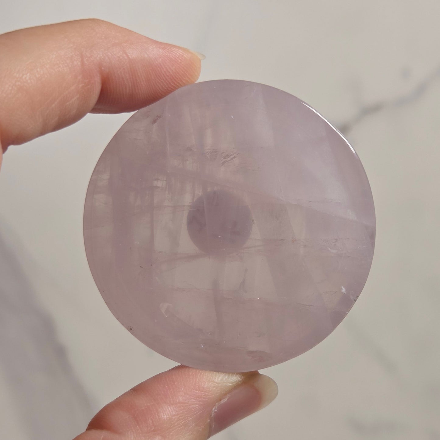 A hand holding a round, translucent light purple stone disc against a light marble-patterned background.