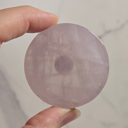 A hand holding a round, translucent light purple stone disc against a light marble-patterned background.