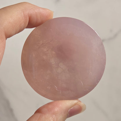 A hand holding a round, polished pink stone or crystal against a light marble-like background.