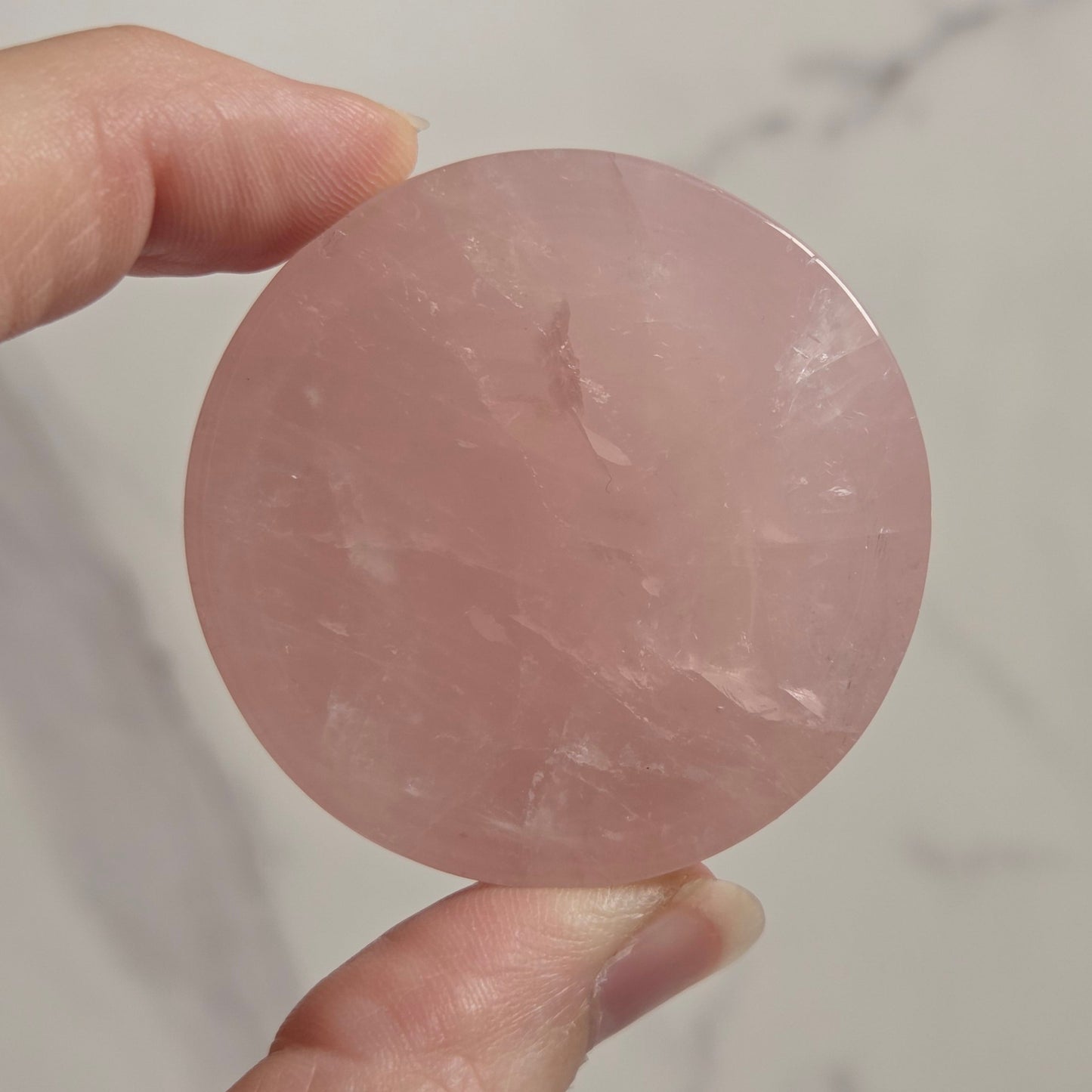 A hand holding a round, polished pink stone or crystal against a light marble-patterned background.