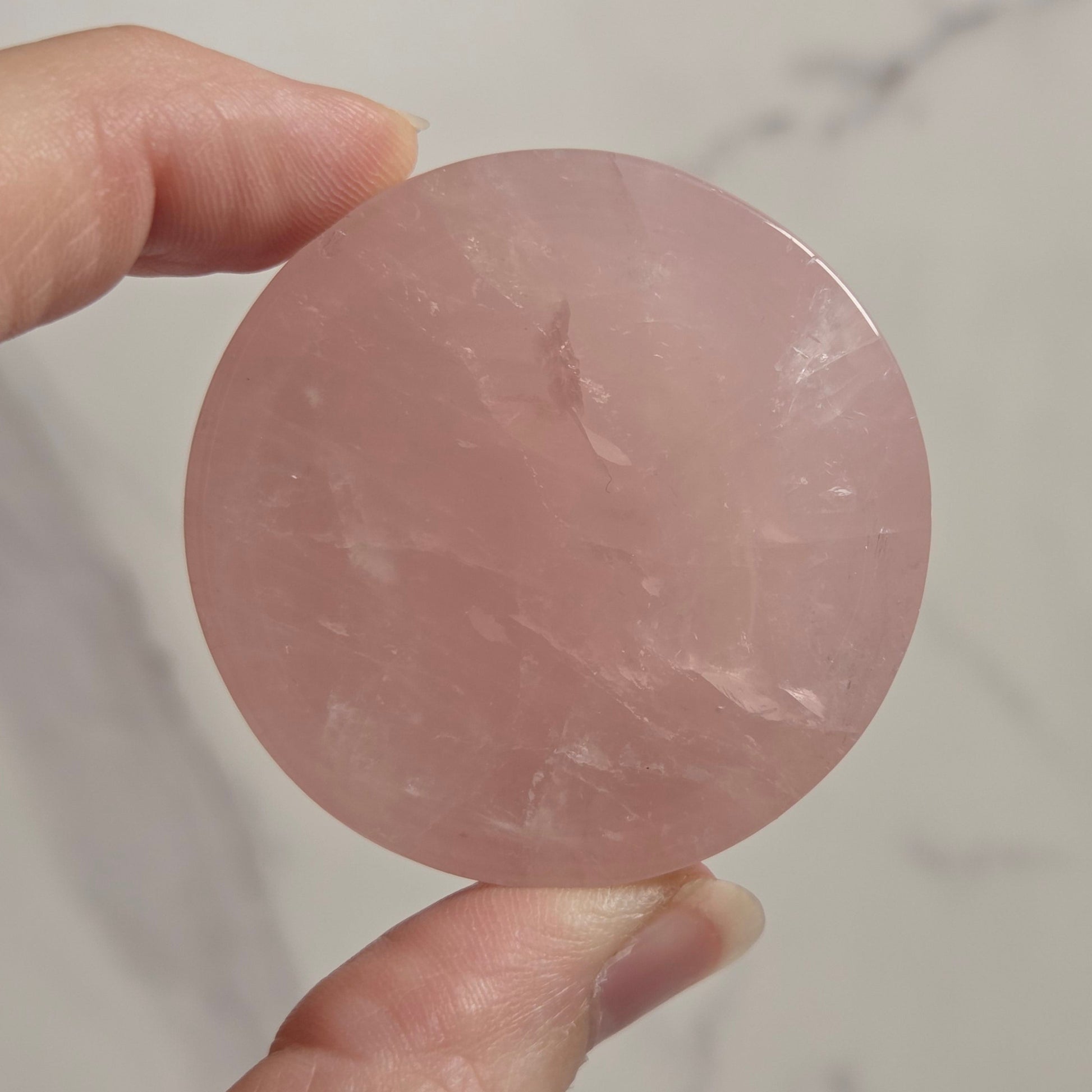 A hand holding a round, polished pink stone or crystal against a light marble-patterned background.