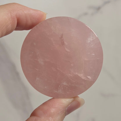A hand holding a round, polished pink stone or crystal against a light marble-patterned background.