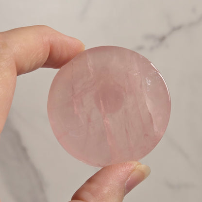 A hand holding a round, polished pink stone or crystal against a light marble-patterned background.
