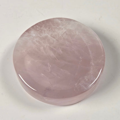 A polished, round rose quartz crystal cabochon with a smooth, glossy finish, resting on a plain off-white surface.