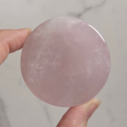 A hand holding a smooth, round, pale pink polished stone or crystal against a light marble-like background.