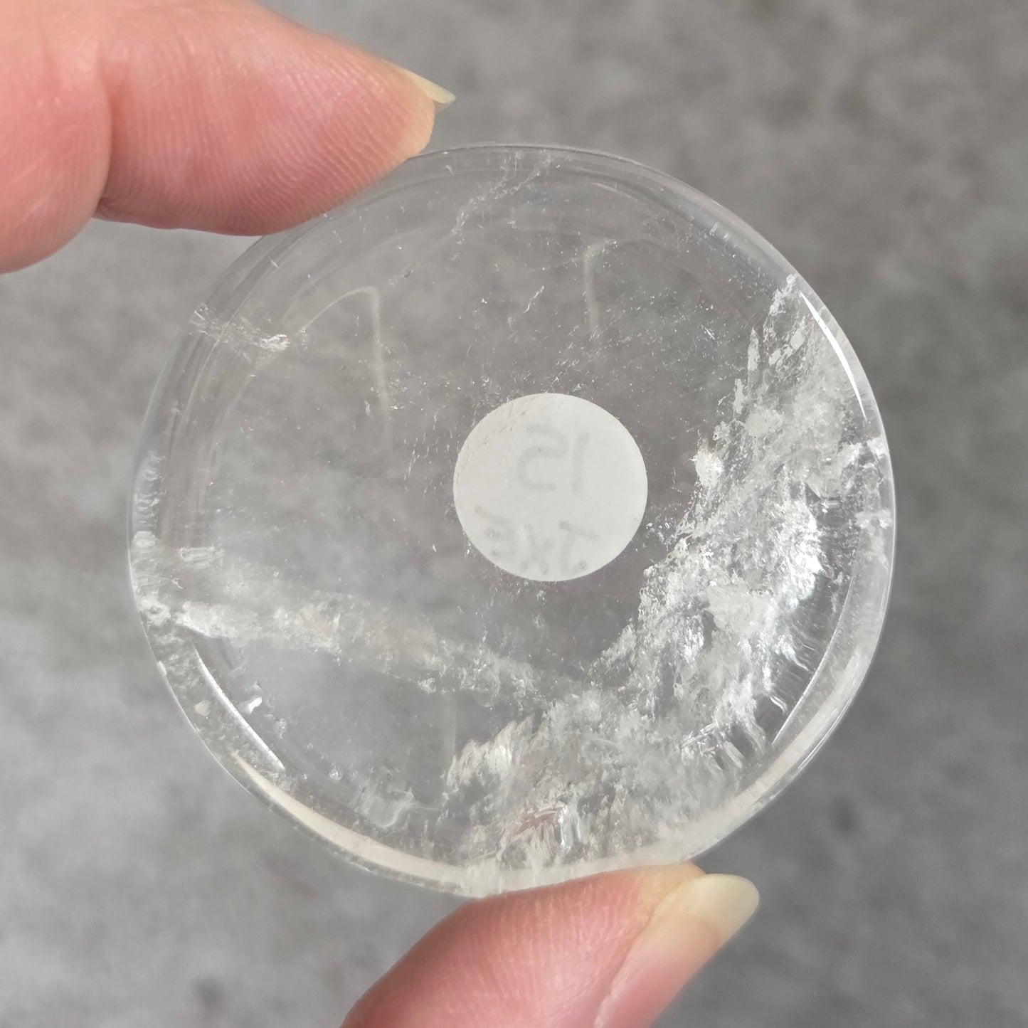 A hand holding a clear, circular plastic or acrylic disc with a small white circular sticker in the center, set against a gray textured background.