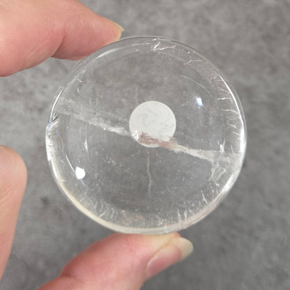 A hand holding a clear, round glass or acrylic button with a white center, shown against a textured gray background.