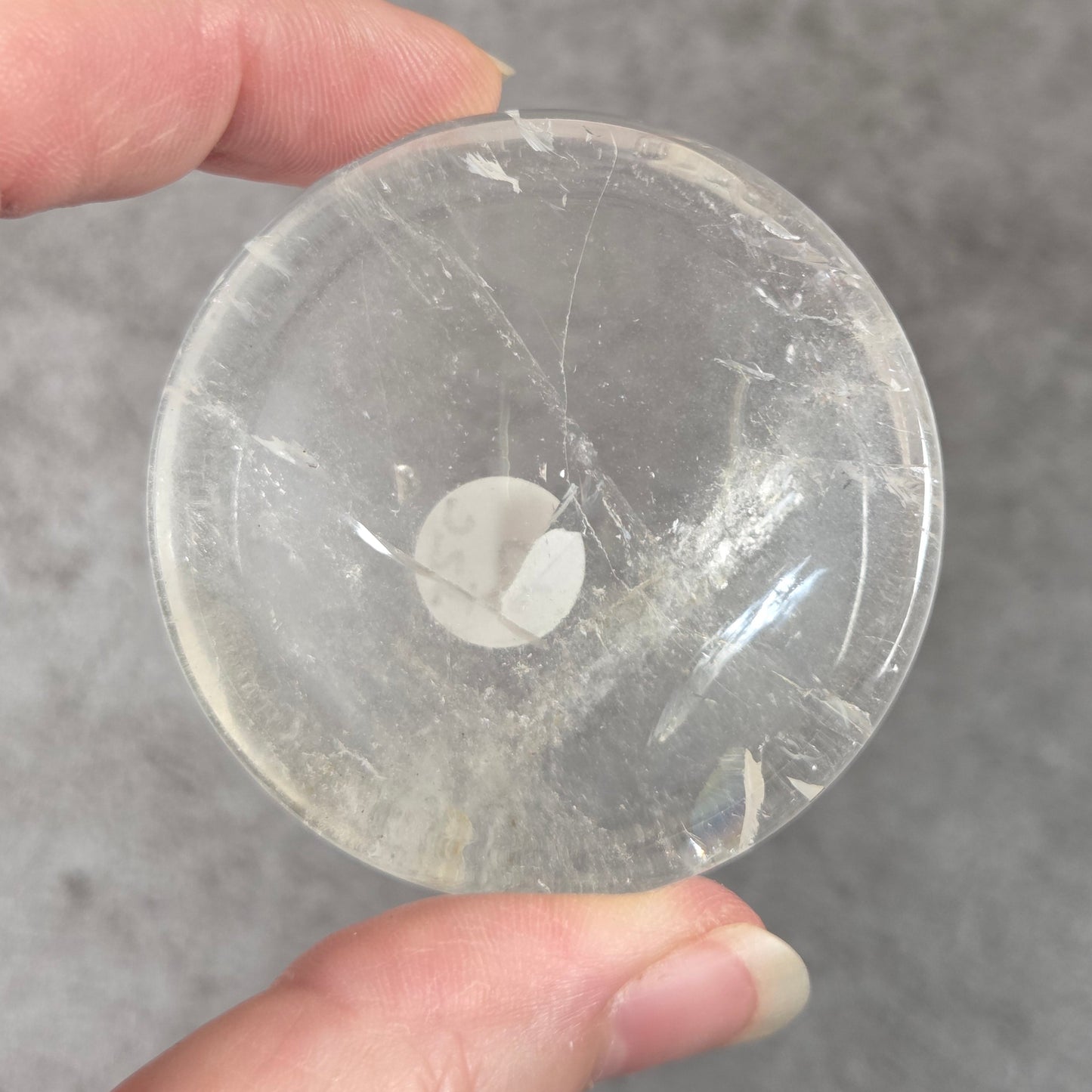 A hand holding a round, clear glass or acrylic button or knob against a gray textured background.