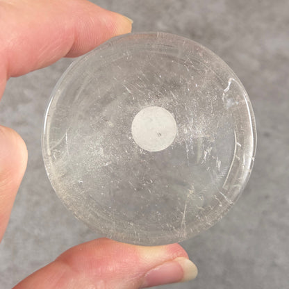 A hand holding a round, clear glass or acrylic coaster with a small white circular center, against a textured gray background.