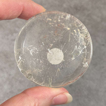 A hand holding a round, clear glass or acrylic dome with visible scratches and a small white circular mark in the center, against a gray textured background.