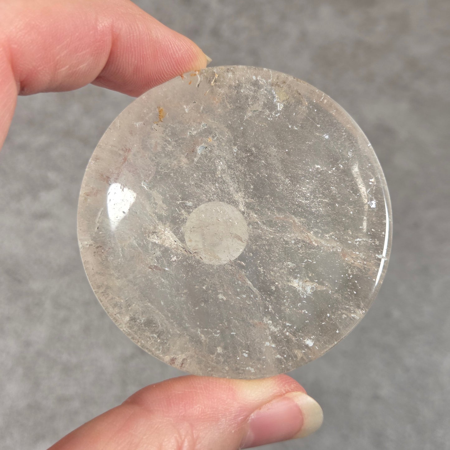 A hand holding a round, polished clear stone disc with natural inclusions, shown against a textured gray background.
