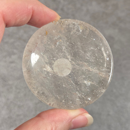 A hand holding a round, polished clear stone disc with natural inclusions, shown against a textured gray background.