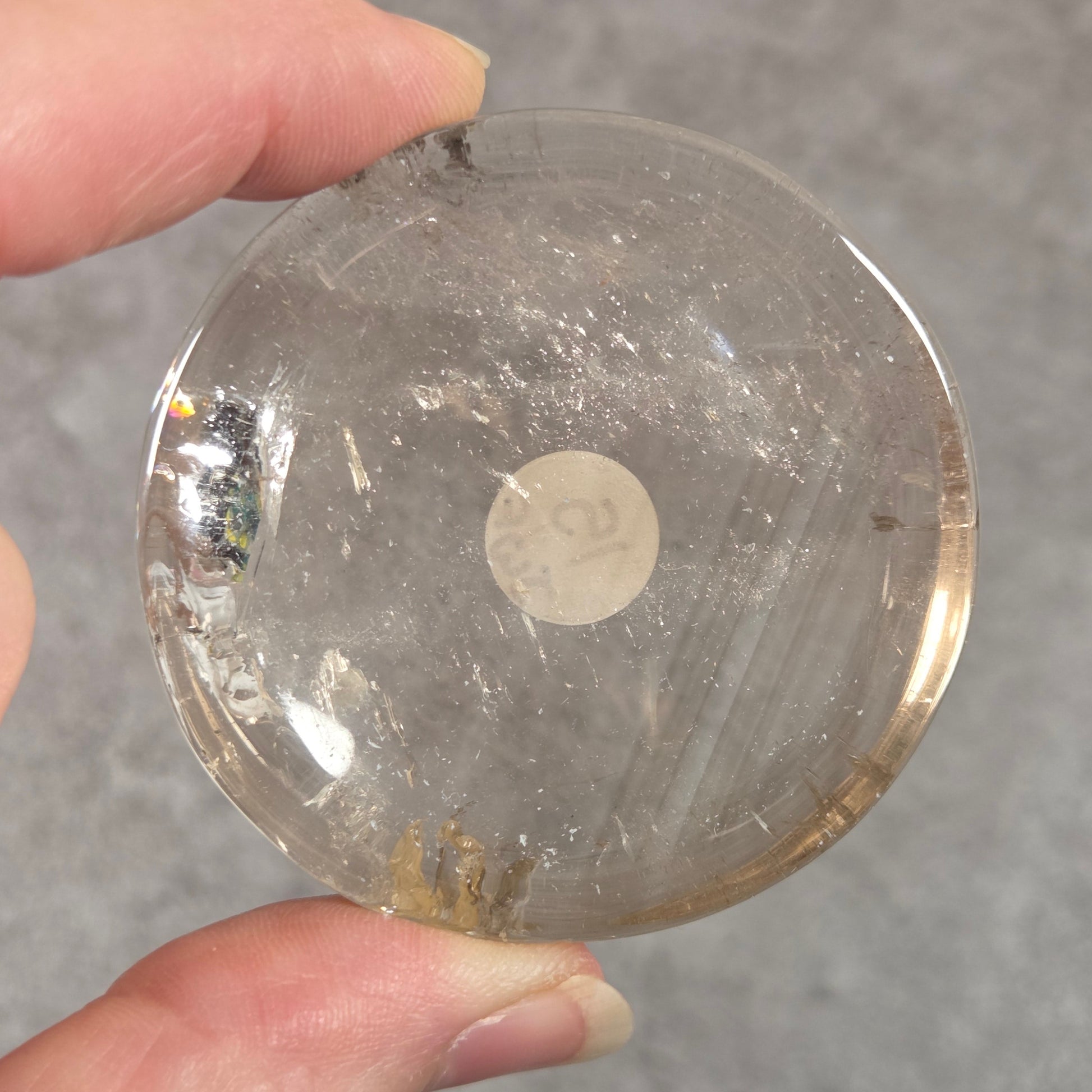 A hand holding a round, clear glass or acrylic coaster with a small white sticker on the bottom, set against a gray textured background.