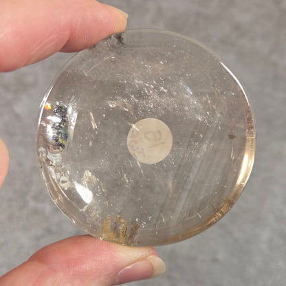 A hand holding a round, clear glass or acrylic coaster with a small white sticker on the bottom, set against a gray textured background.