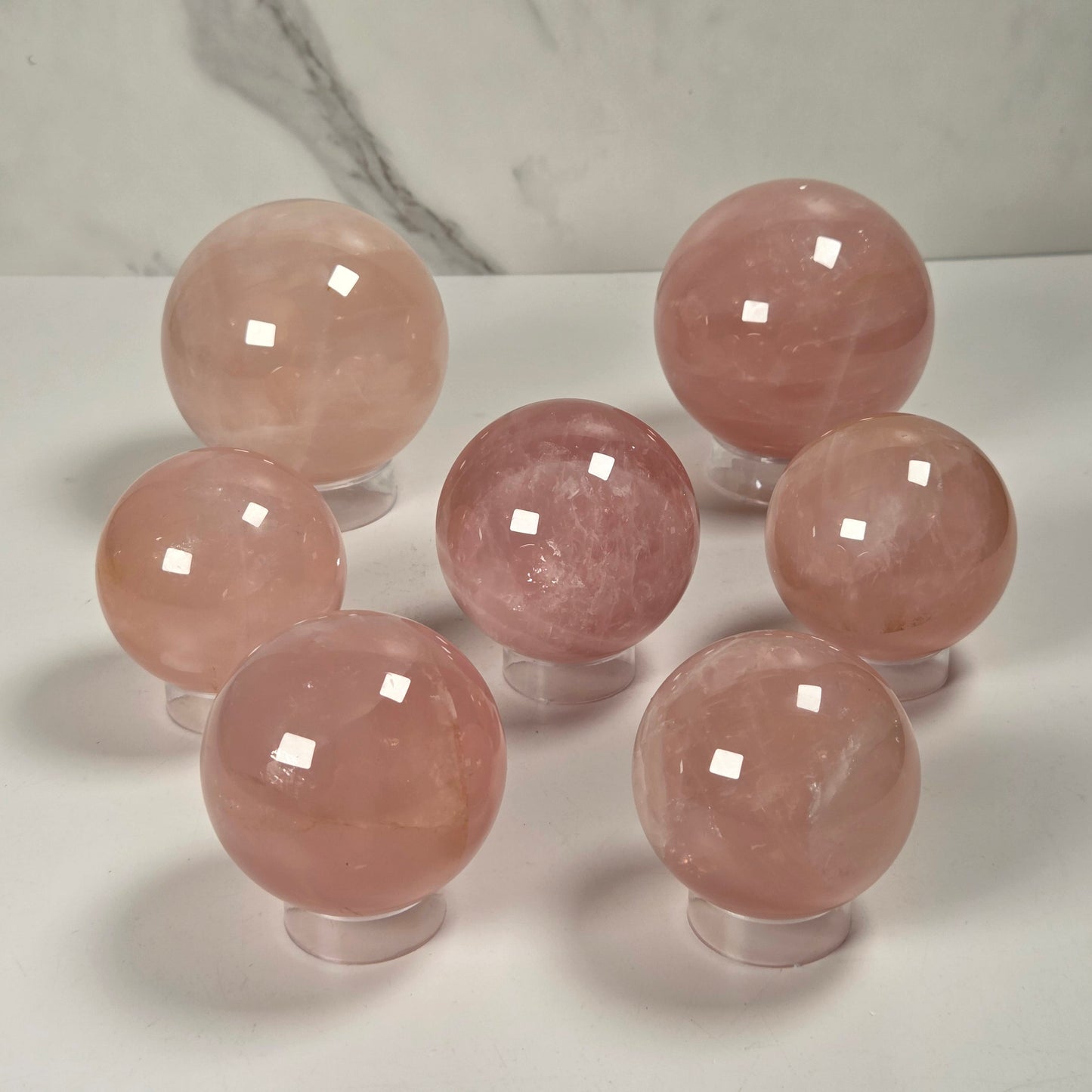 Seven polished pink crystal spheres displayed on clear stands against a white surface with a light gray marble-patterned background.