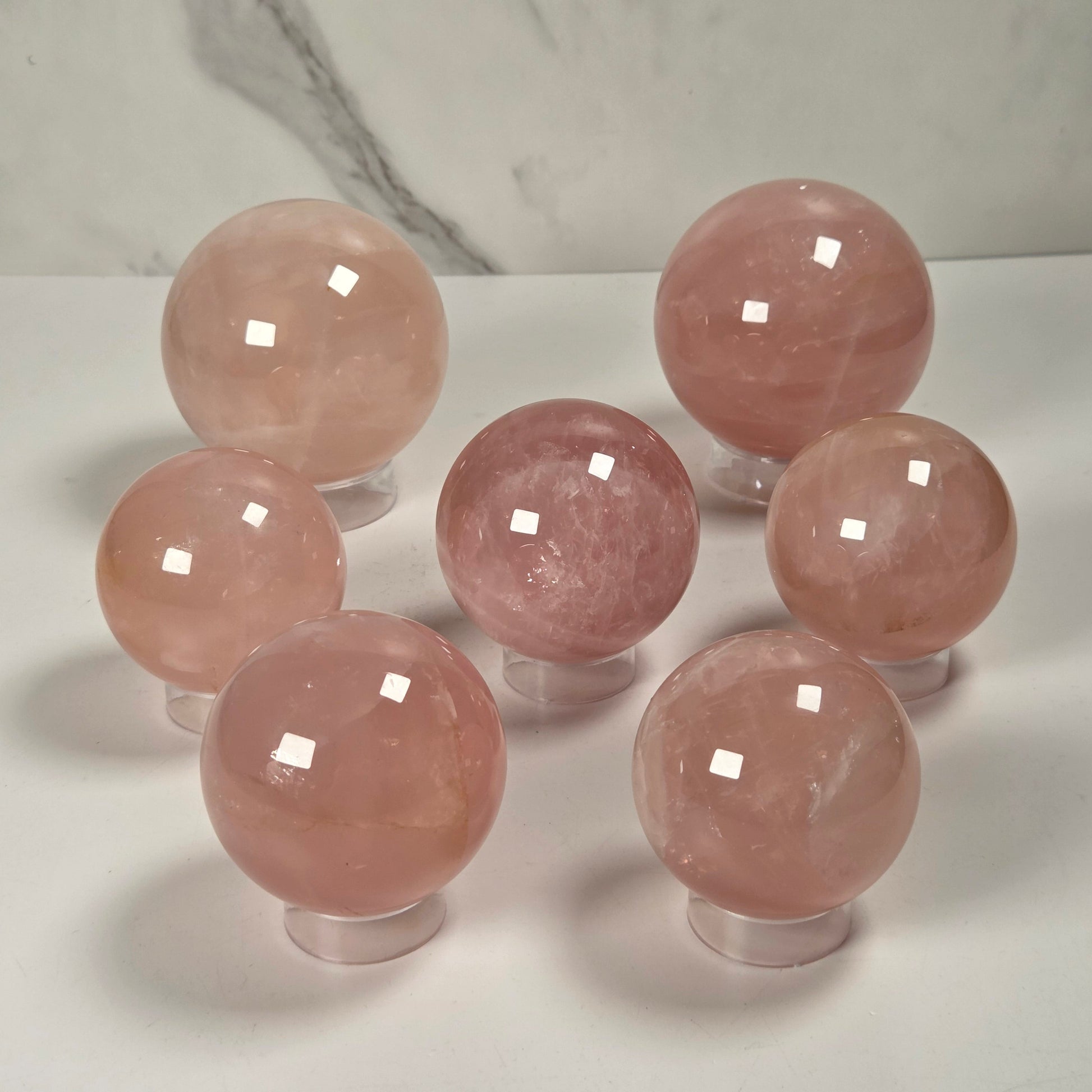 Seven polished pink crystal spheres displayed on clear stands against a white surface with a light gray marble-patterned background.