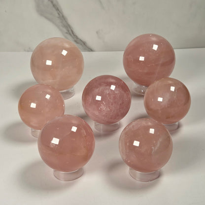 Seven polished pink crystal spheres displayed on clear stands against a white surface with a light gray marble-patterned background.