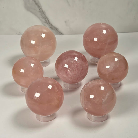 Seven polished pink crystal spheres displayed on clear stands against a white surface with a light gray marble-patterned background.