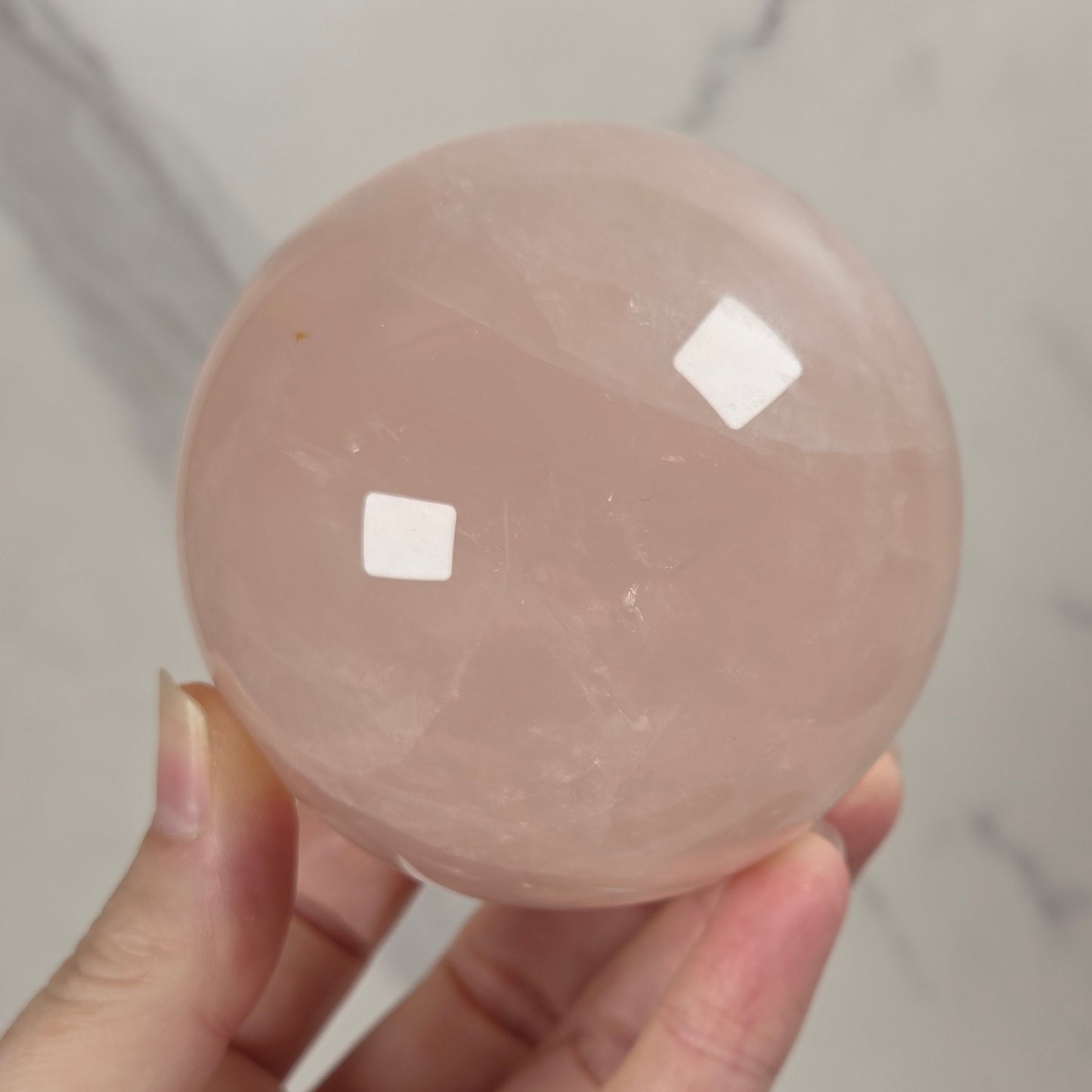 A hand holding a smooth, polished pink rose quartz crystal sphere against a light gray marble background.
