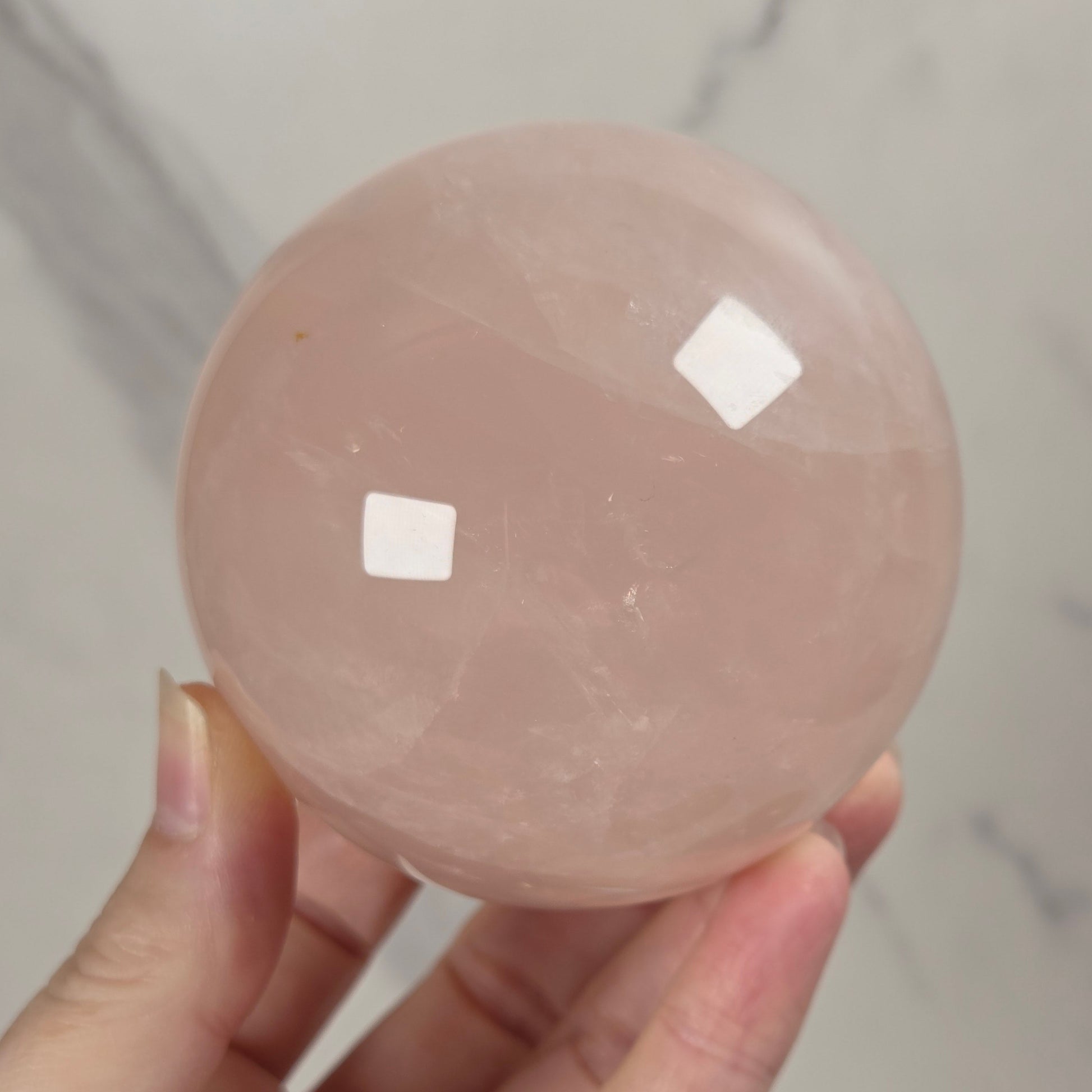 A hand holding a smooth, polished pink rose quartz crystal sphere against a light gray marble background.