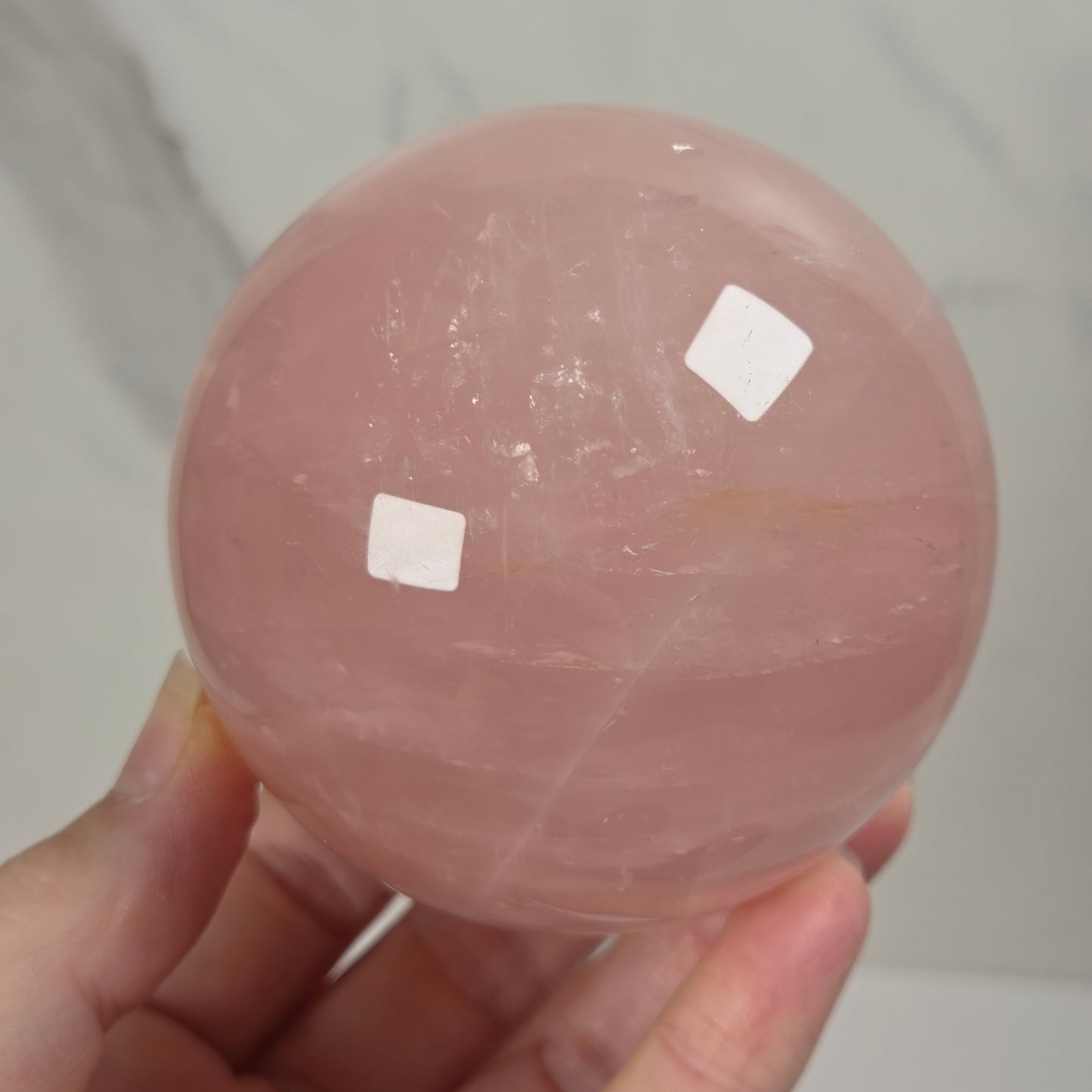 A hand holding a smooth, polished pink rose quartz crystal sphere against a light gray marble 