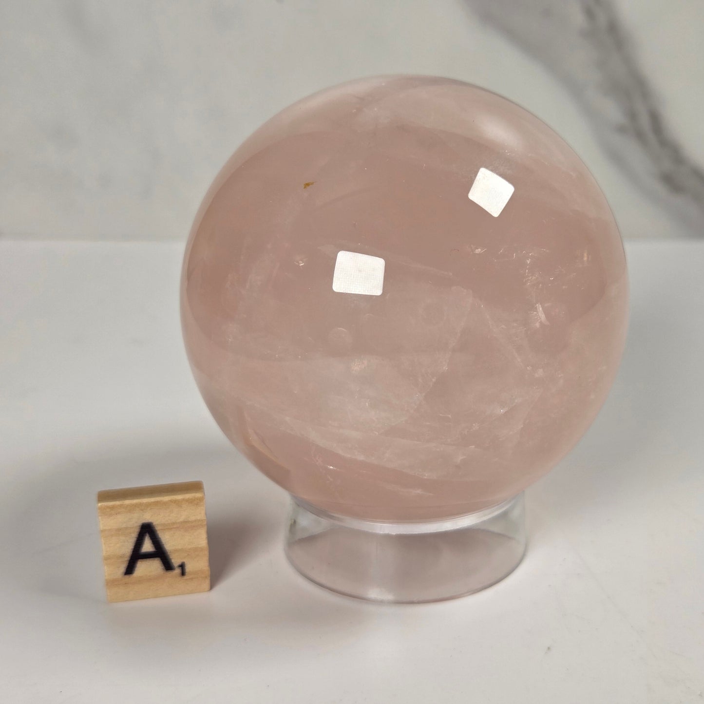 A large smooth pink quartz crystal sphere on a clear acrylic stand, with a small wooden letter tile showing 'A1' beside it on a white surface against a light marble-patterned background.