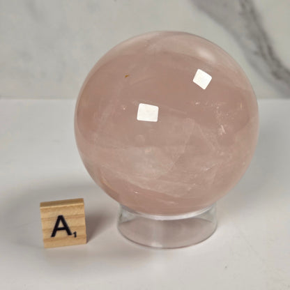 A large smooth pink quartz crystal sphere on a clear acrylic stand, with a small wooden letter tile showing 'A1' beside it on a white surface against a light marble-patterned background.
