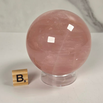A polished pink rose quartz crystal sphere on a clear acrylic stand, shown next to a small wooden Scrabble tile on a white surface with a light gray marble-patterned background.