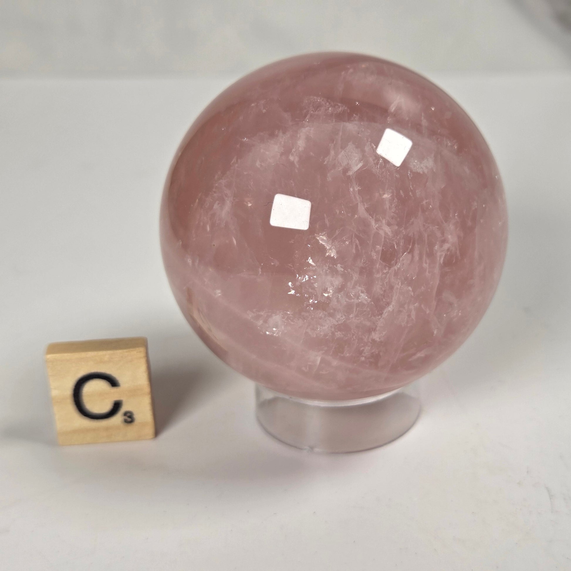 A polished pink rose quartz crystal sphere on a clear acrylic stand, shown next to a small wooden Scrabble tile spelling 'C3' on a light gray marble surface.
