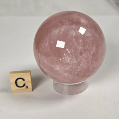 A polished pink rose quartz crystal sphere on a clear acrylic stand, shown next to a small wooden Scrabble tile spelling 'C3' on a light gray marble surface.