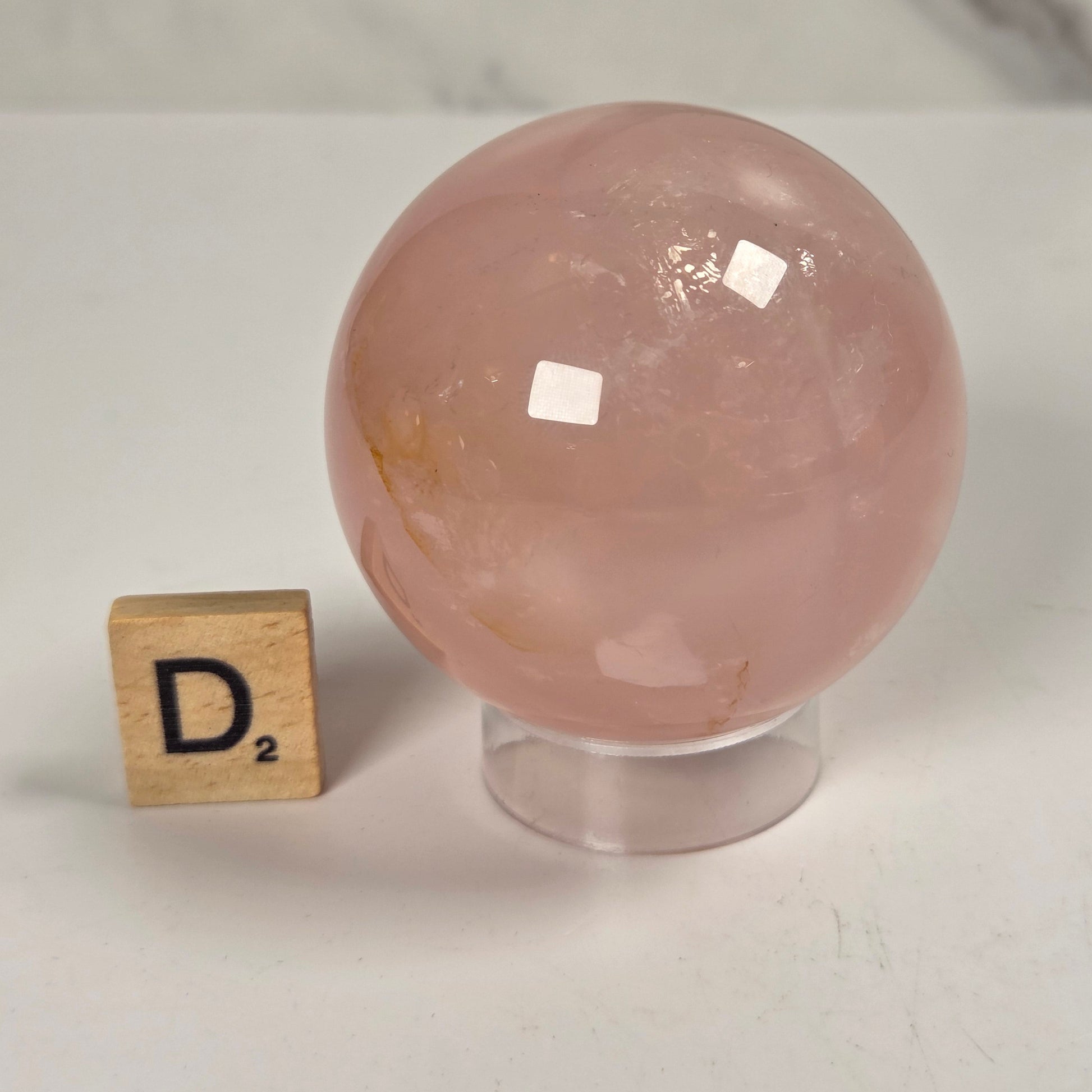 A polished pink rose quartz crystal sphere on a clear acrylic stand, shown next to a small wooden Scrabble tile spelling 'D²' on a light marble surface.