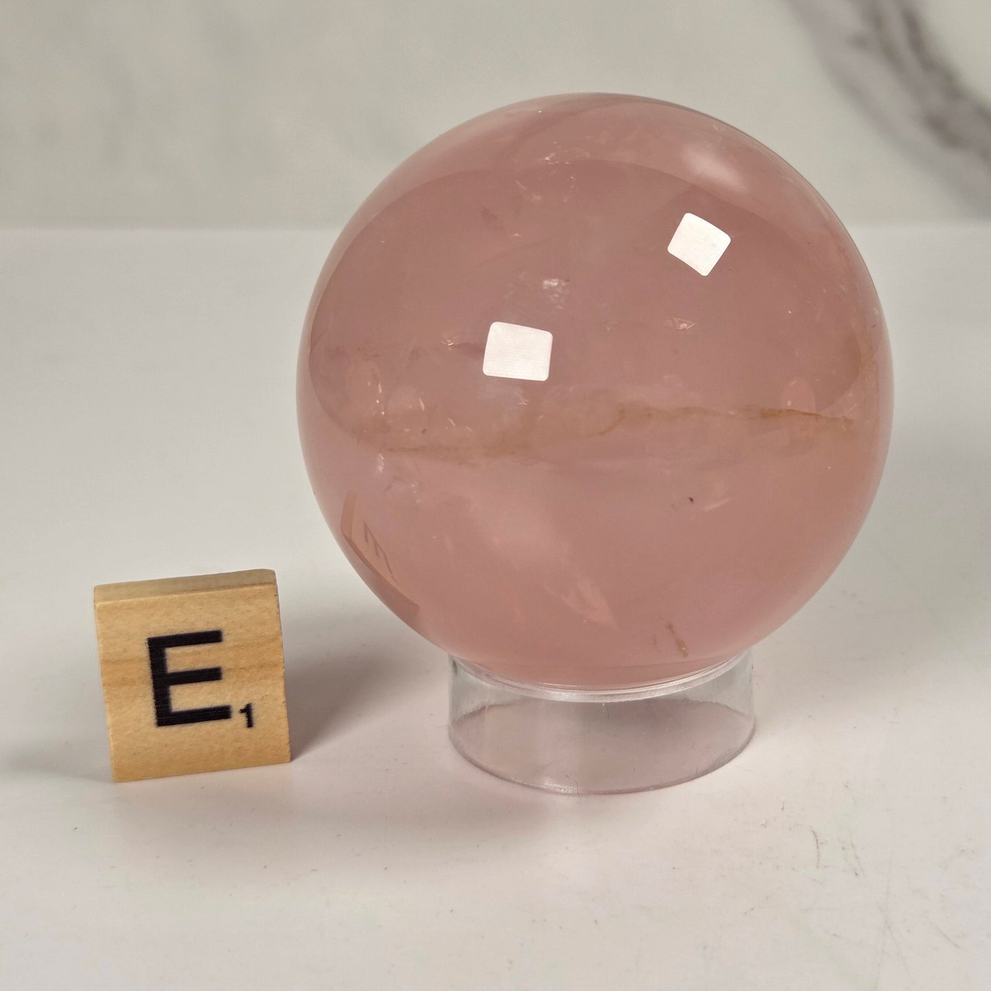 A polished pink rose quartz crystal sphere on a clear acrylic stand, shown next to a small wooden Scrabble tile spelling 'E²' on a light marble surface.