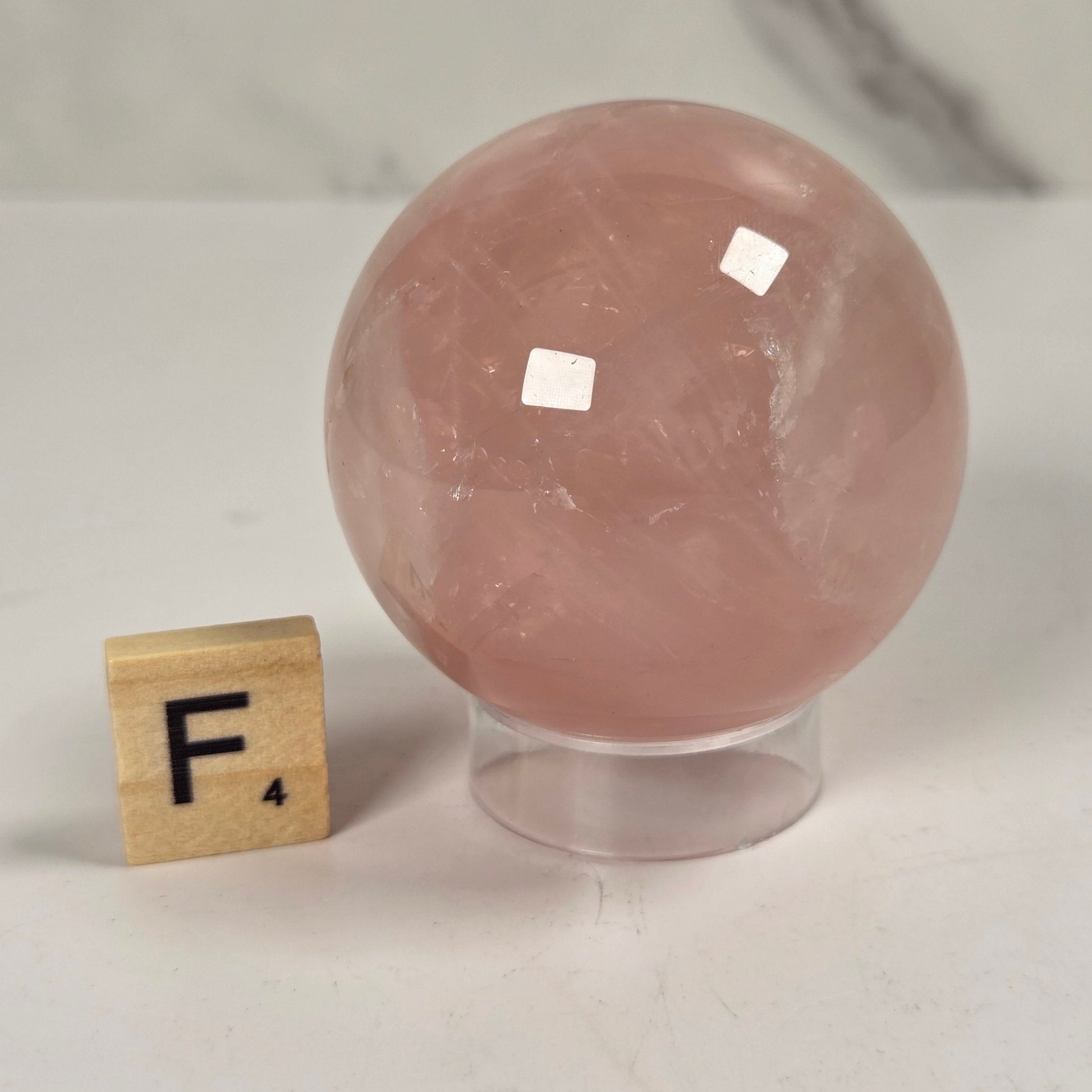 A pink rose quartz crystal sphere on a clear acrylic display stand, shown next to a wooden Scrabble tile with the letter F on a light marble surface.