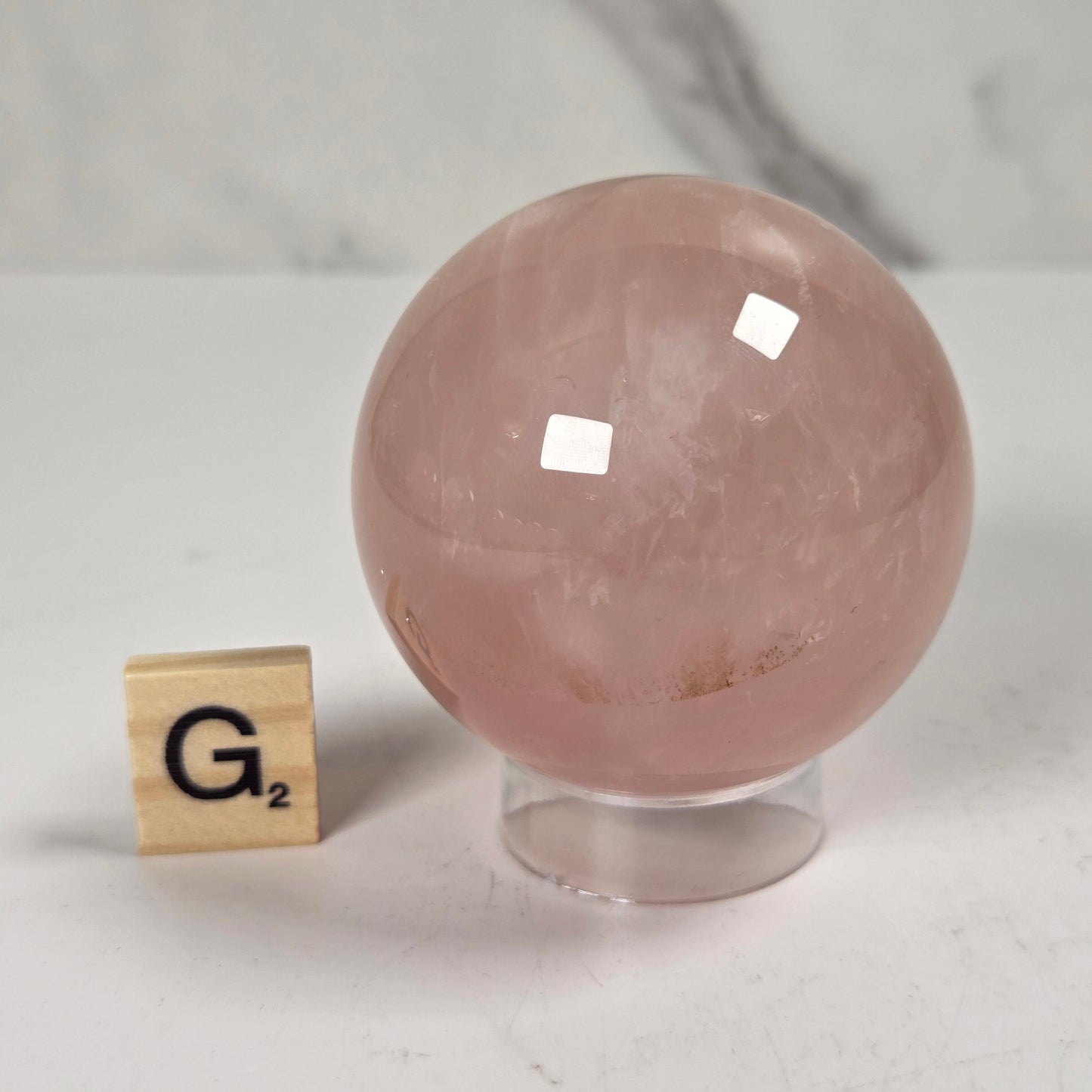 A polished pink quartz crystal sphere on a clear acrylic stand, shown next to a wooden Scrabble tile with the letter G on a white marble surface.