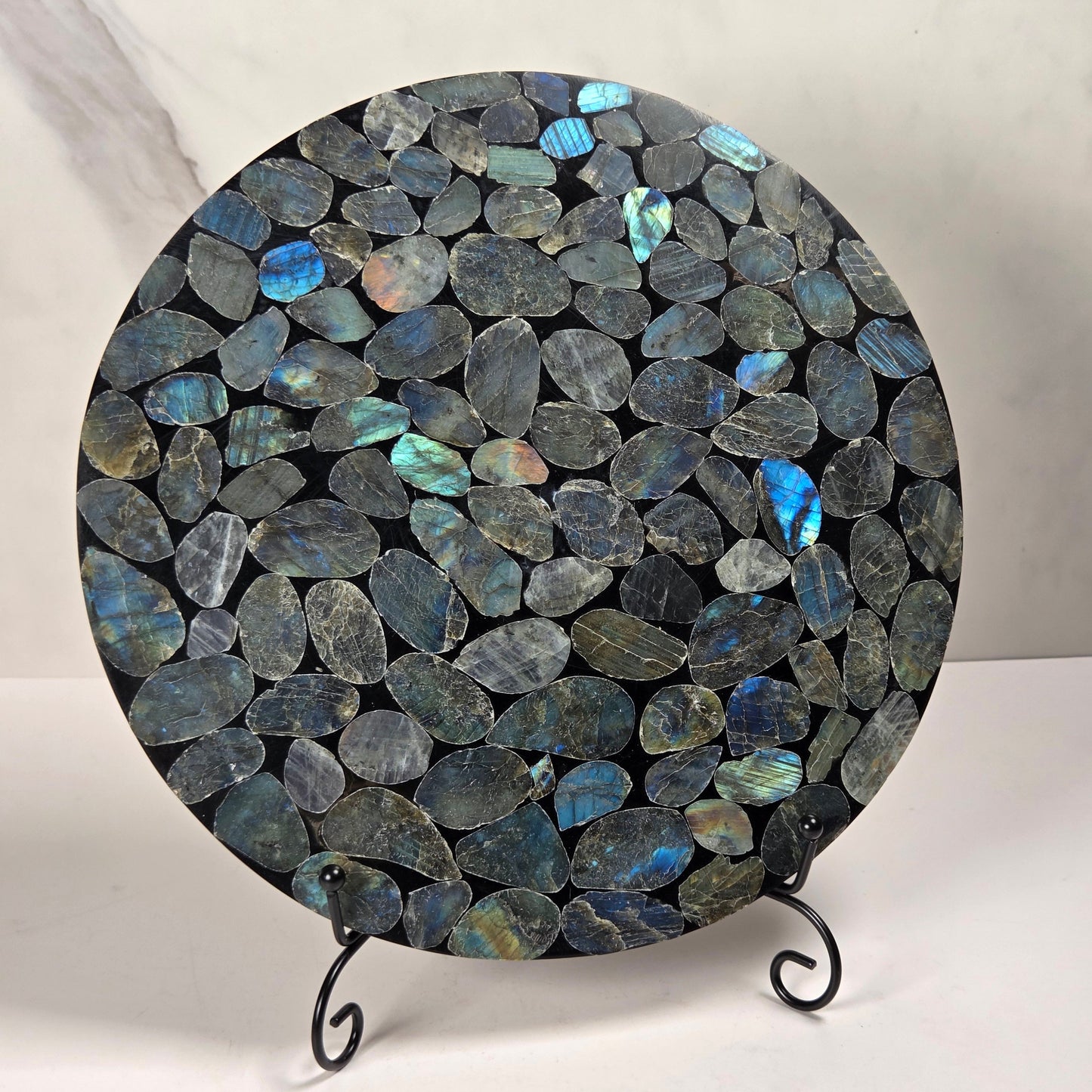 A round decorative board or tray covered in polished irregular labradorite stones with blue, green, and gray flashes, displayed on a small black metal stand against a light marble-patterned background.