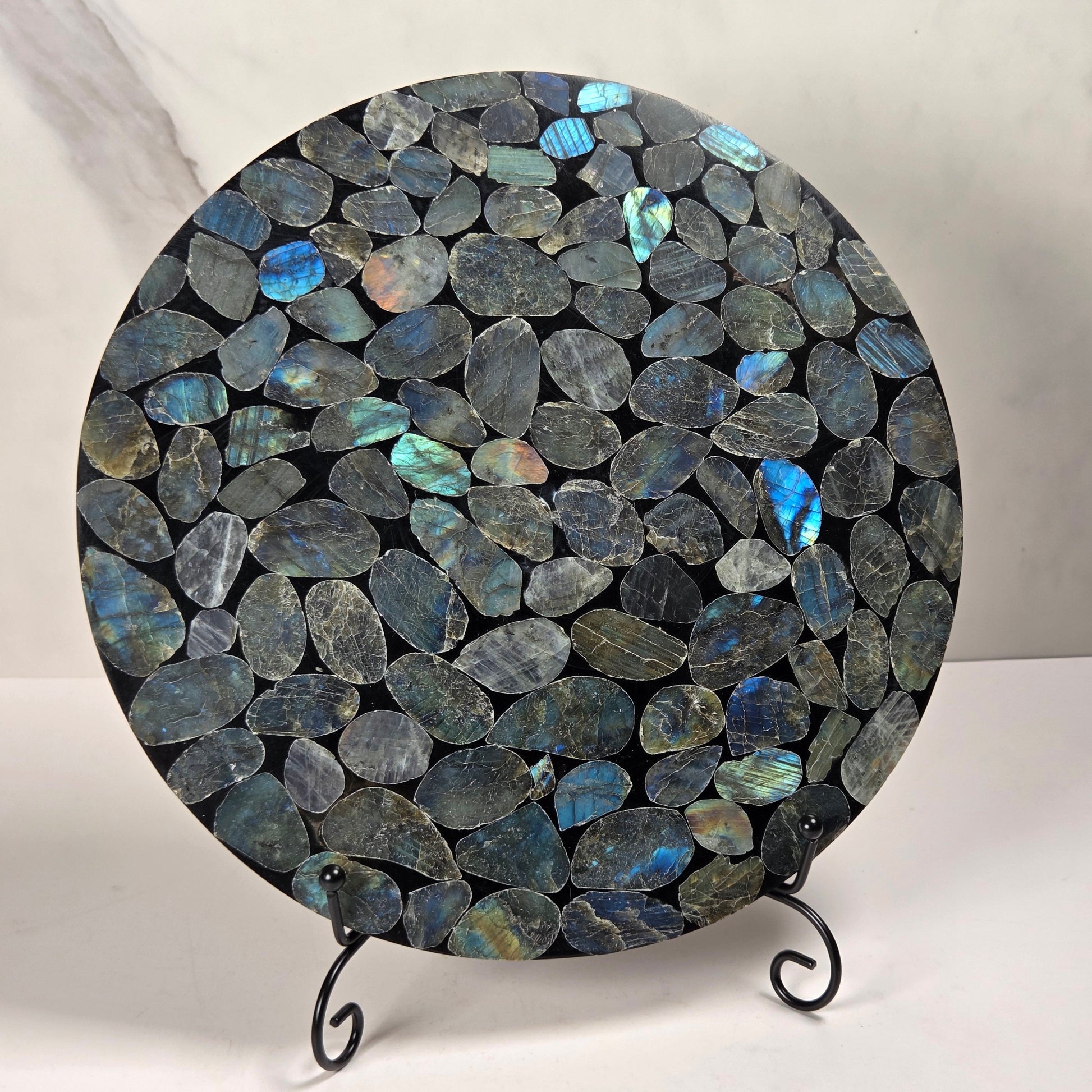 A round decorative board or tray covered in polished irregular labradorite stones with blue, green, and gray flashes, displayed on a small black metal stand against a light marble-patterned background.